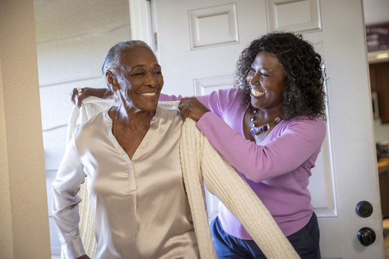 A woman assists an older woman in putting on a sweater, showcasing a moment of care and support between them.