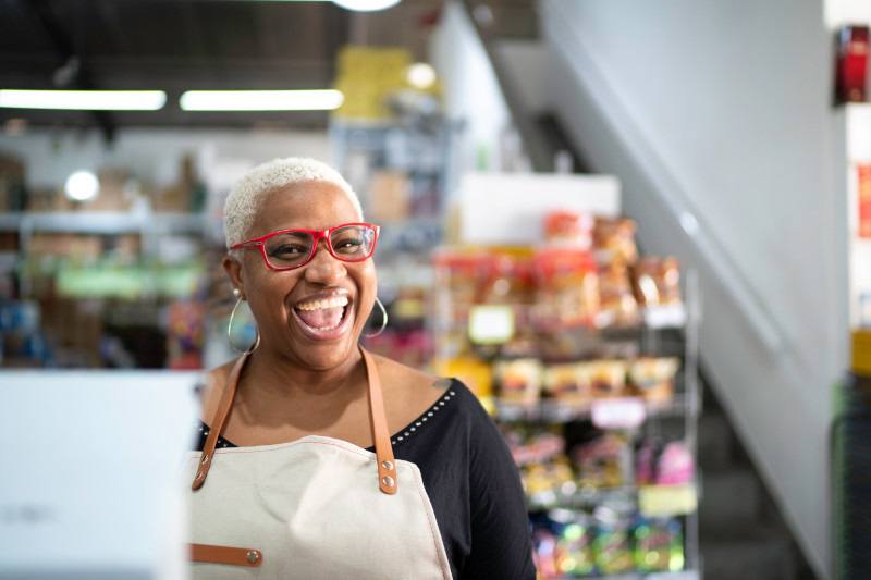 A smiling woman wearing an apron stands in a store, surrounded by shelves filled with various products.